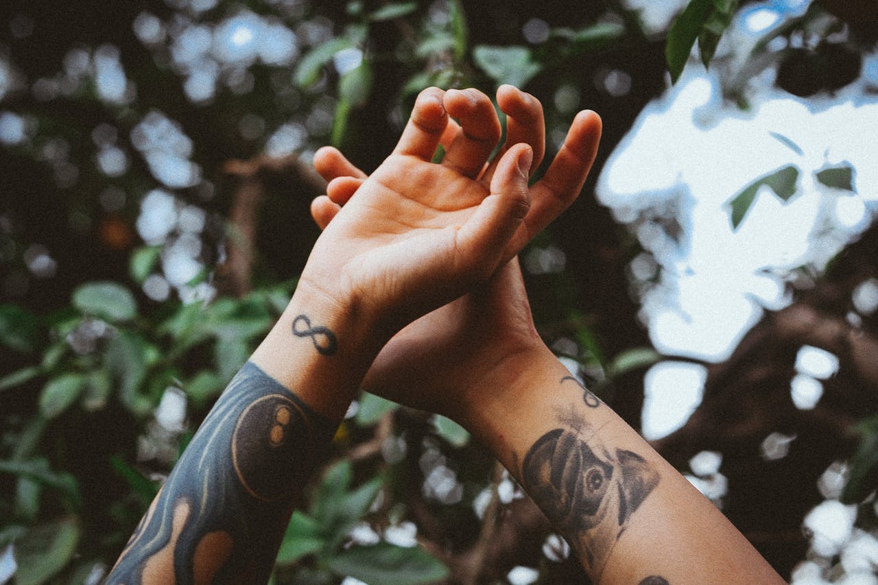Close-up of tattooed hands reaching out against a blurry natural backdrop.